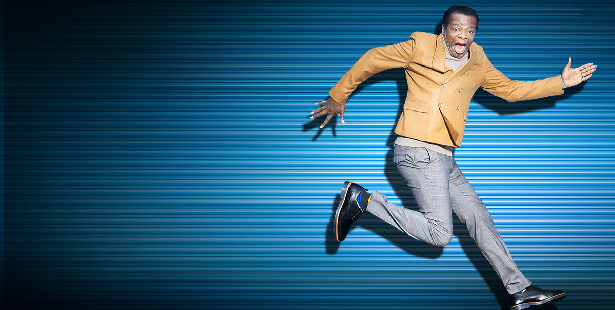 Image description: Stephen K Amos jumps in front of a blue wall with funny look on his face